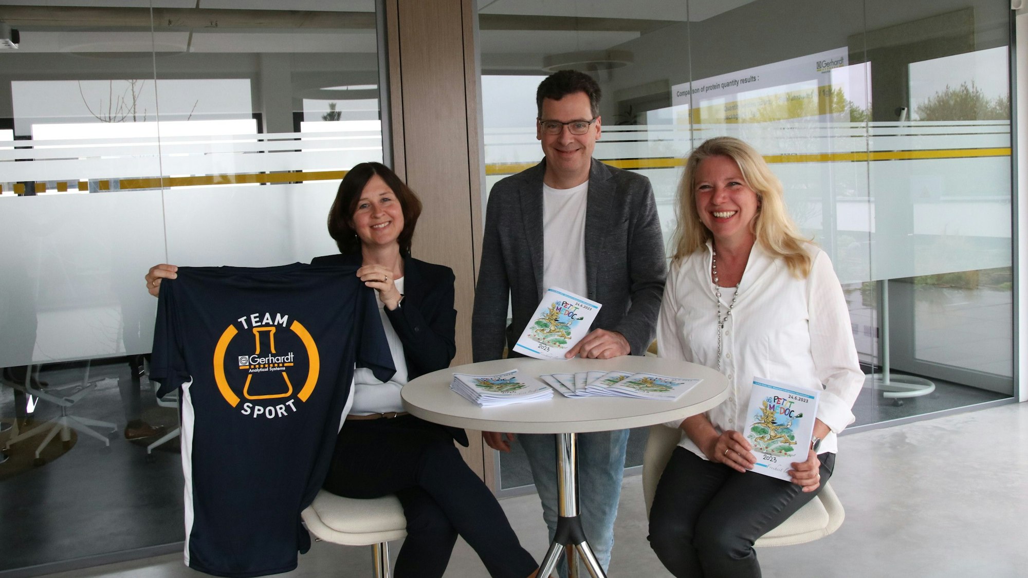 Ein Mann und zwei Frauen stehen beziehungsweise sitzen an einem hohen Tisch und zeigen Flyer mit Informationen zum „Petit Medoc“ sowie ein T-Shirt.