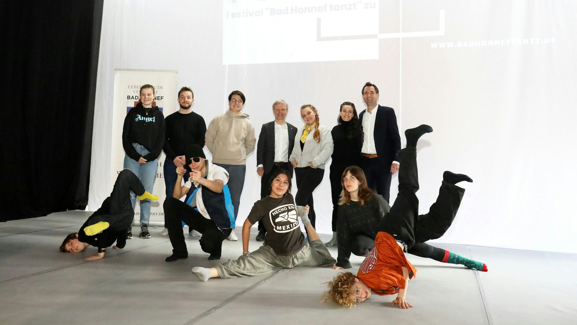 Eine Gruppe von Menschen steht im Ratssaal vor einer weißen Wand. Einige Kinder und Jugendliche zeigen Tanz-Elemente, wohl aus Rap oder Breakdance.