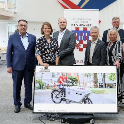 Acht Frauen und Männer stehen hinter einem großformatigen Monitor, auf dem eine Fahrerin mit einem Lastenrad zu sehen ist, mit dem das „Rhein-Kaufhaus“ Waren auslieferte.