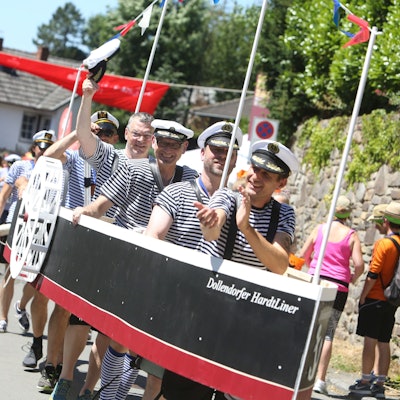 Junge Männer, die blau-weiße T-Shirts und Kapitänsmützen tragen, stecken in einem großen Modellschiff, das am Bug den Namen „Dollendorfer Hardtliner“ trägt.