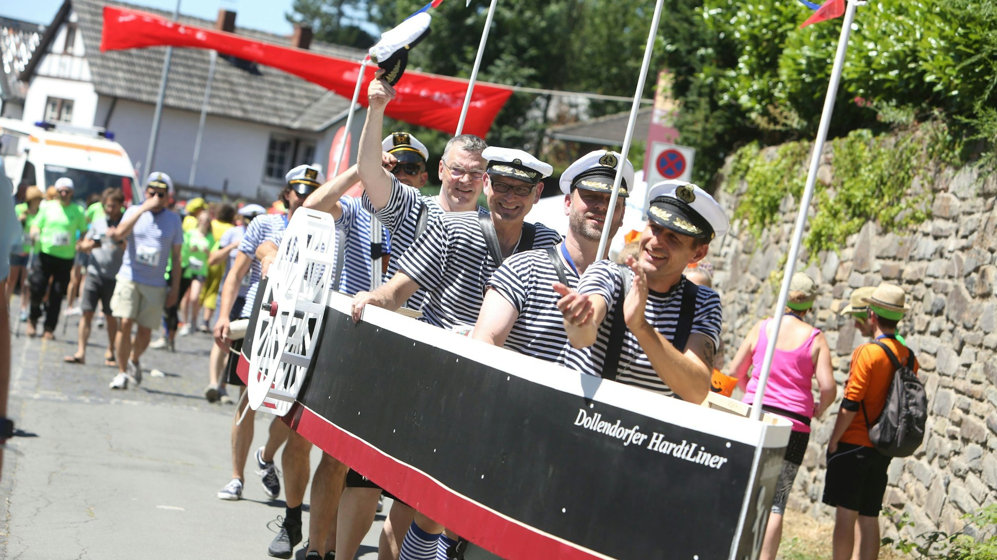 Junge Männer, die blau-weiße T-Shirts und Kapitänsmützen tragen, stecken in einem großen Modellschiff, das am Bug den Namen „Dollendorfer Hardtliner“ trägt.