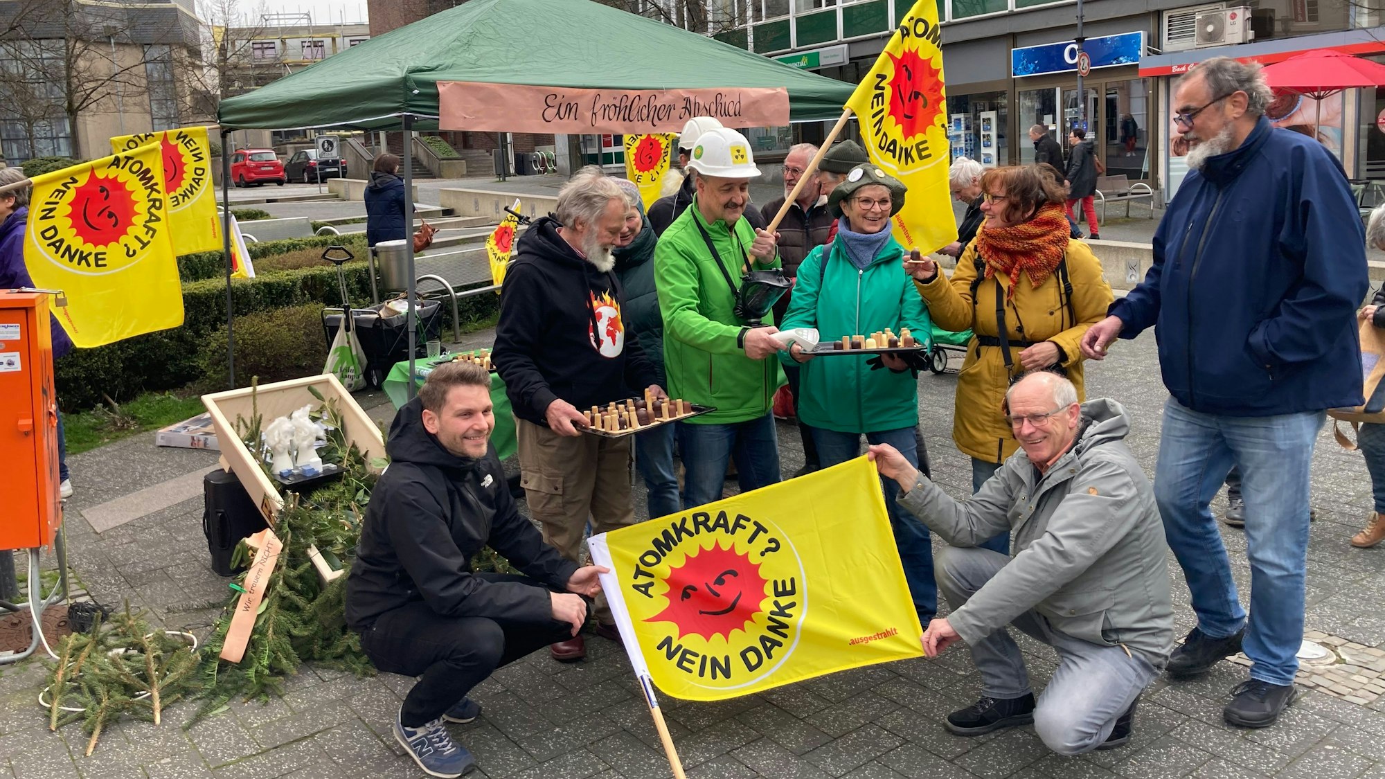 Mehrere Personen stehen und knien, mit gelben Fahnen, auf denen „Atomkraft? Nein Danke!“ steht, in einer Fußgängerzone.