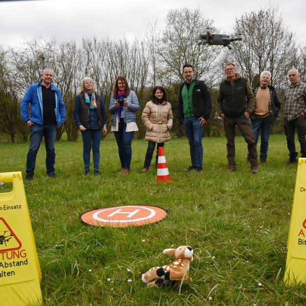 Mehrere Leute stehen auf einer Wiese in einer Reihe. Einer fliegt mit einer Drohne über ein im Gras liegendes Stoff-Rehkitz. Daneben stehen zwei gelbe Warnschilder mit der Aufschrift: „Drohne im Einsatz. Achtung, bitte Abstand halten“.