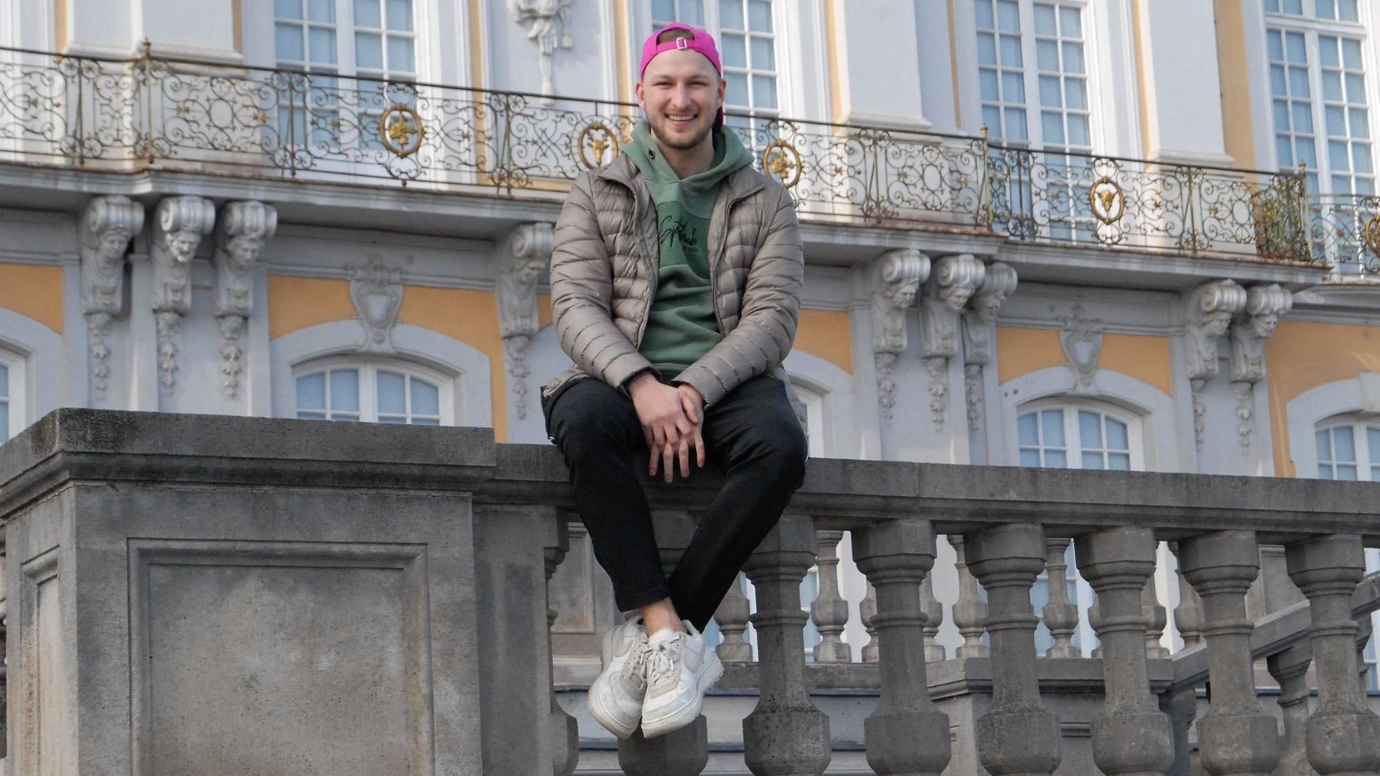 Auf dem Fot ist DSDS-Teilnehmer David Leischik vor dem Schloss in Brühl zu sehen. Er sitzt auf einem Geländer.