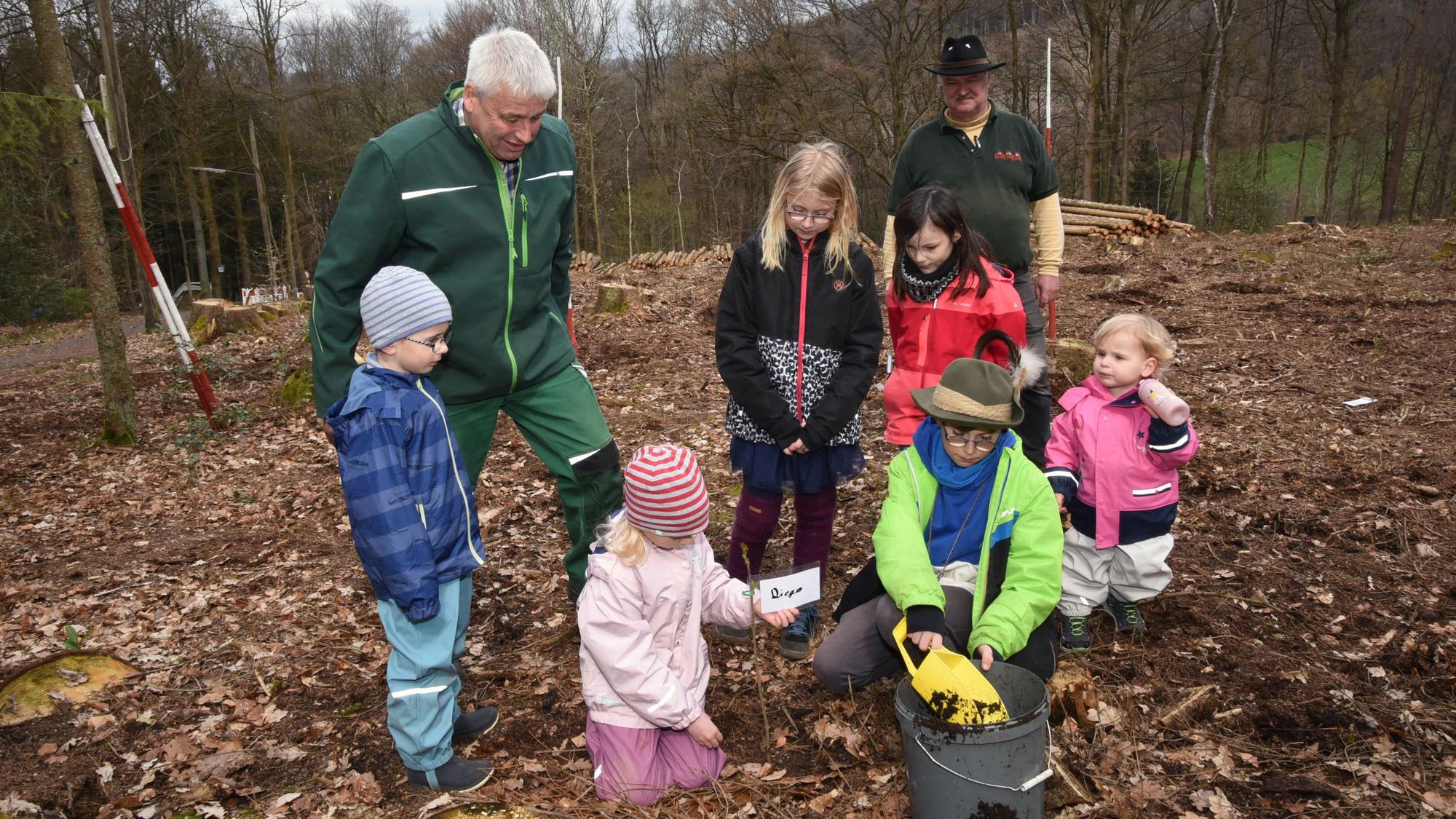 Mehrere Kinder pflanzen unter Anleitung eines älteren Mannes in grünem Overall kleine Setzlinge.
