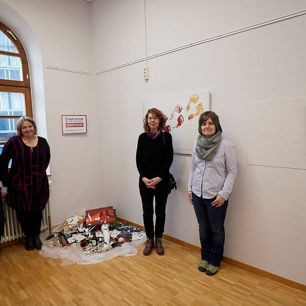 Bilder in der Galerie Eifel Kunst werden gezeigt sowie wie vier Frauen neben einem Kunstwerk über hausliche Gewalt.