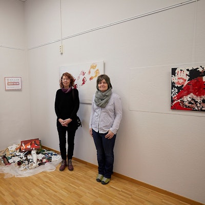 Bilder in der Galerie Eifel Kunst werden gezeigt sowie wie vier Frauen neben einem Kunstwerk über hausliche Gewalt.