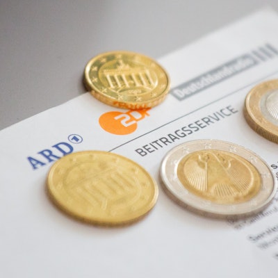 Euromünzen liegen auf einem Anschreiben zur Anmeldung für die Rundfunkgebühren, neben den Logo von ARD, ZDF und Deutschlandfunk.