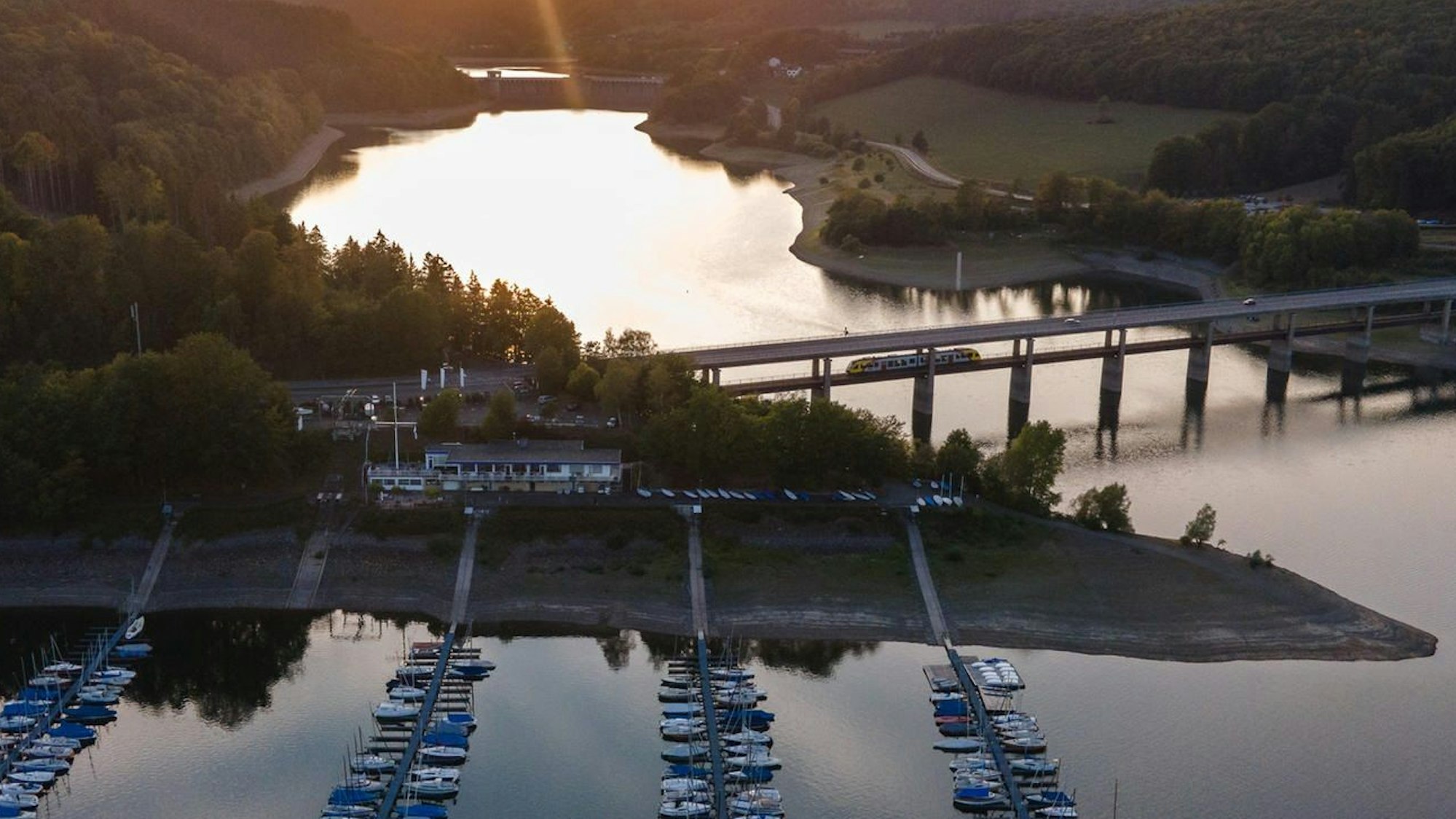 Die Sonne geht am Biggesee hinter einem Segelhafen und einer Brücke unter.