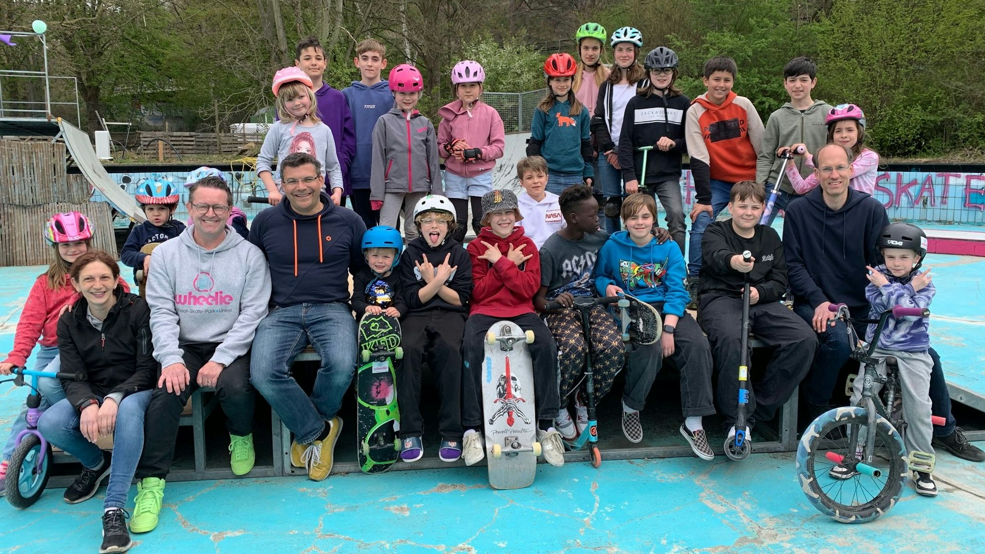 Eine Gruppe von Kindern, Jugendlichen und Erwachsenen haben sich mit Rollern, Skateboards und Fahrrädern zum Gruppenbild postiert. Viele Kinder tragen Fahrradhelme.