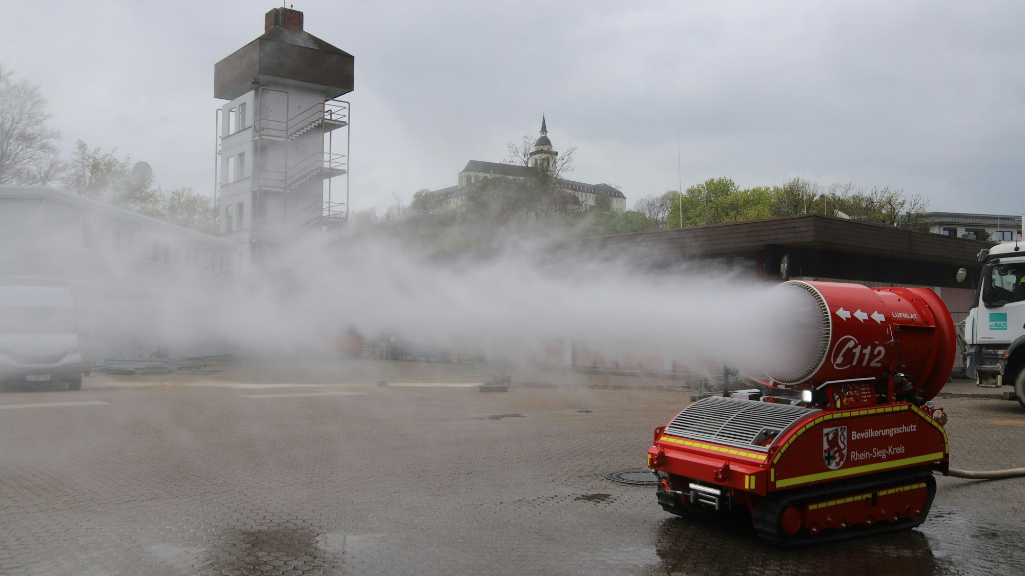 Auf einem kettenangetriebenen flachen Fahrzeug versprüht eine große Düse Wassernebel. Im Hintergrund erhebt sich die Siegburger Abtei auf dem Michaelsberg.