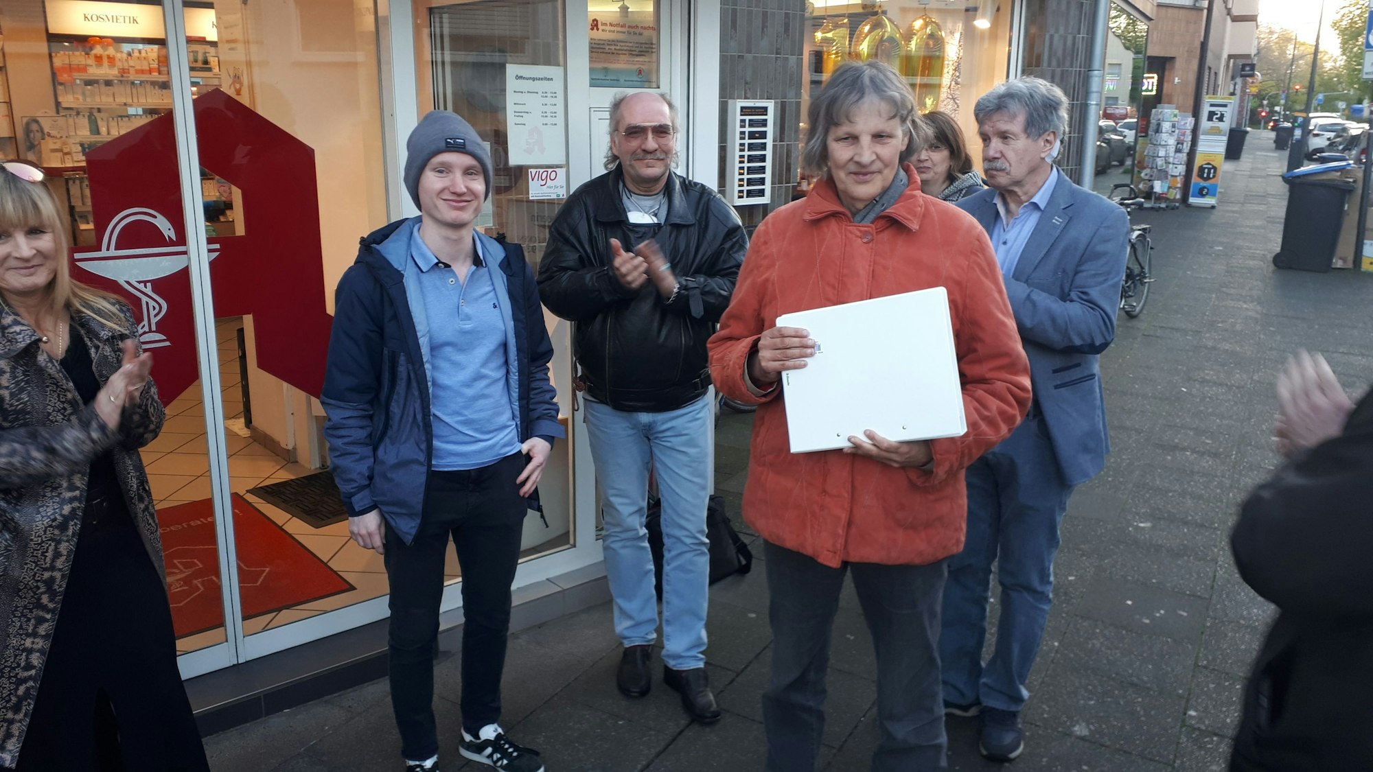 Ein junger Mann steht neben einer älteren Frau, die einen Ordner in der Hand hält. Daneben sind weitere Personen zu sehen. Alle stehen auf einem Bürgersteig vor einer Apotheke.