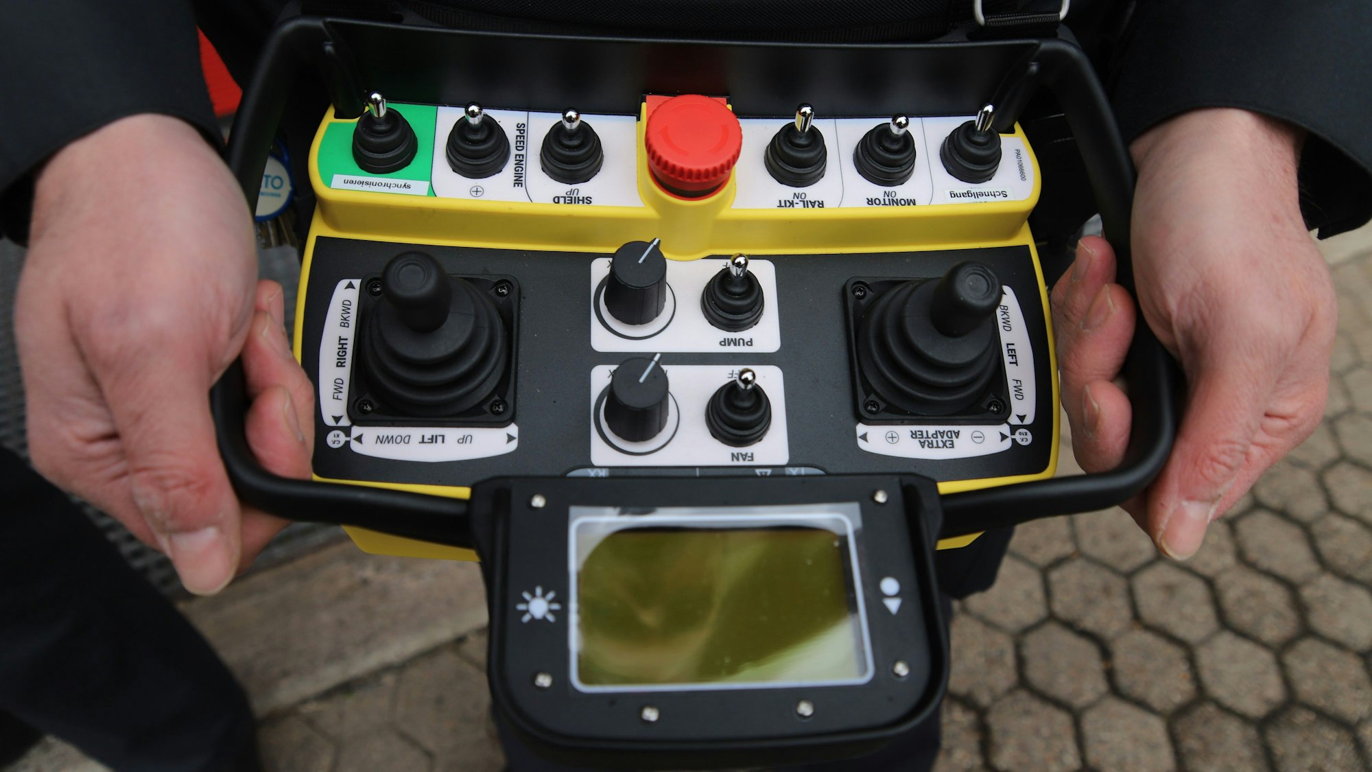 Zwei Hände halten eine Fernsteuerung mit einem Sichtfeld, vielen kleinen Hebeln und einem roten Knopf.