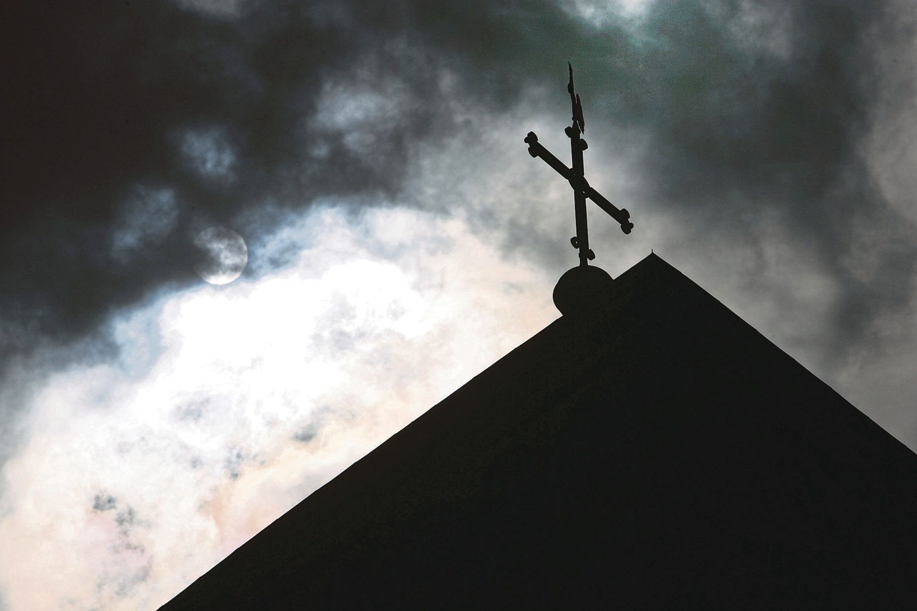Im Gegenlicht und vor wolkenverhangenem Himmel ist die Kirchturmspitze des Doms mit Kreuz zu sehen.