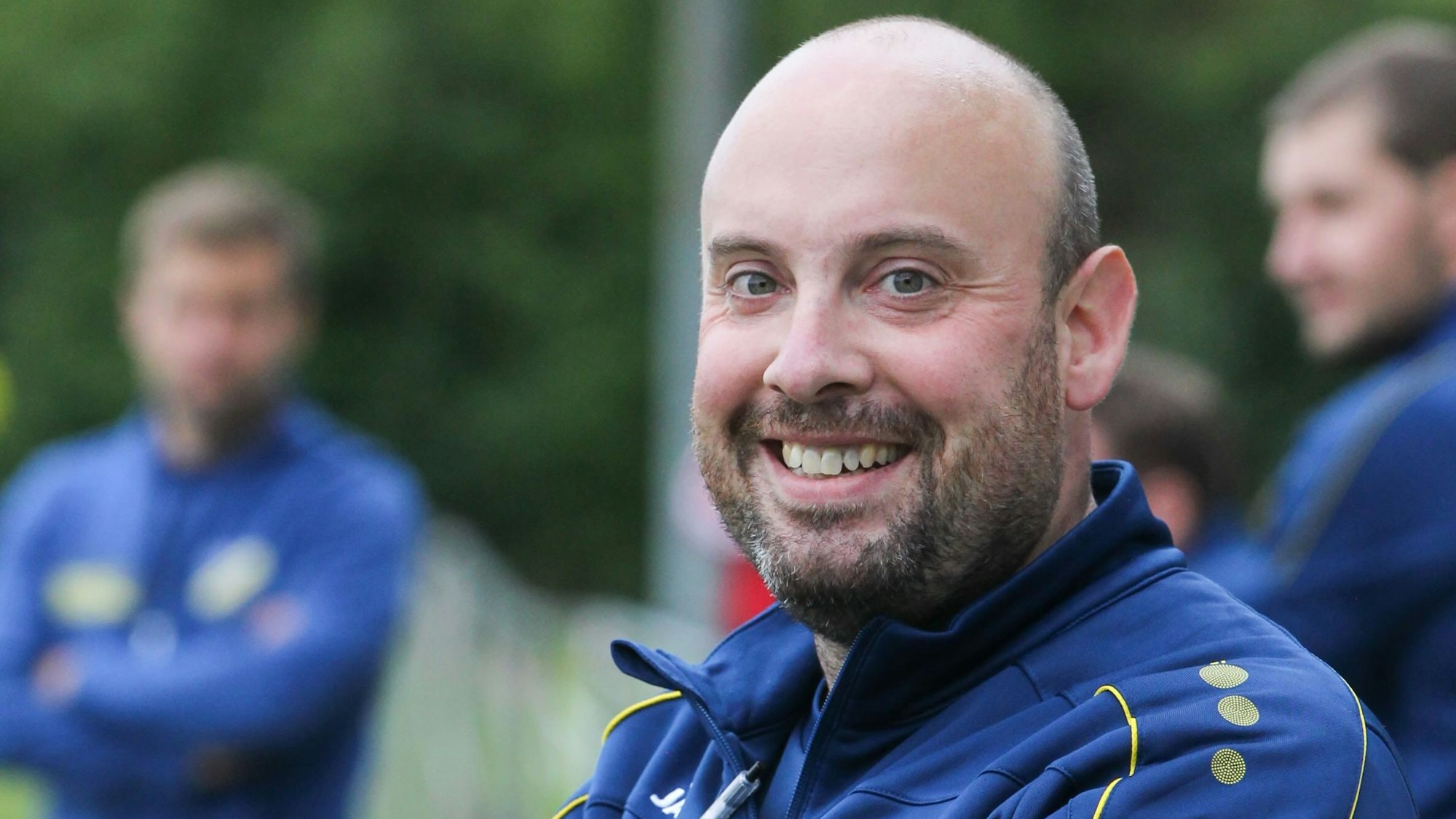 Trainer André Knips lächelt in die Kamera. Er trägt eine blaue Trainingsjacke. Im Hintergrund stehen zwei weitere Männer.