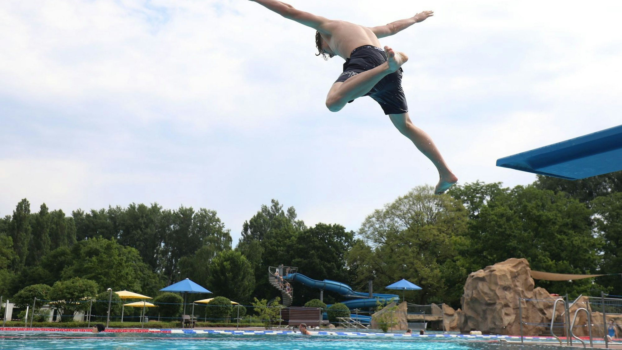 Ein junger Mann springt mit ausgestreckten Armen von einem Sprungbrett ins Bad Honnefer Freibad, Im Hintergrund ist die große blaue Wasserrutsche zu sehen.