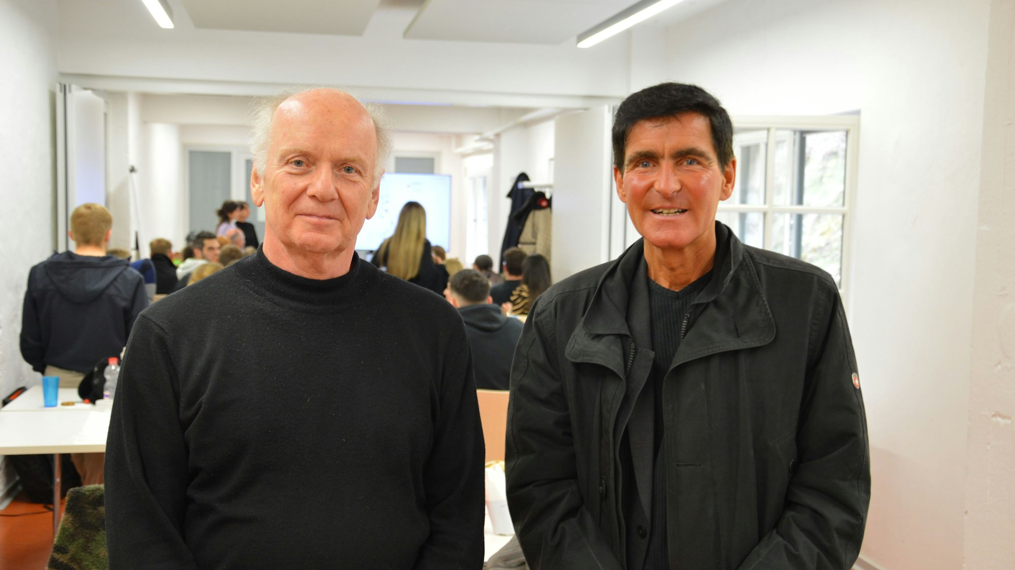 Zwei Männer stehen in einem Raum, im Hintergrund sind Personen zu erkennen.