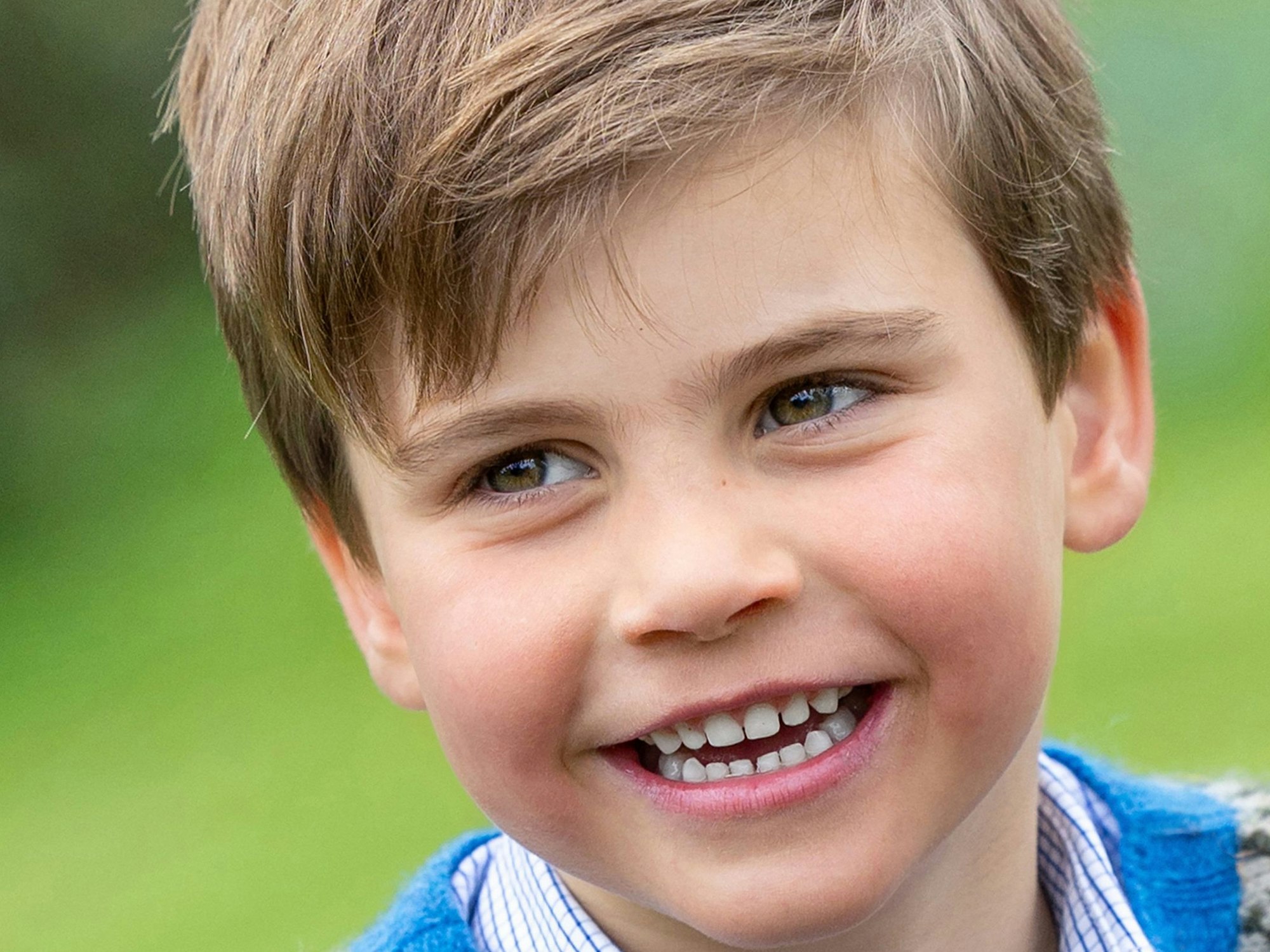 Prinz Louis trägt einen blauen Pullover, darunter ein kariertes Hemd. Er lächelt in die Kamera.