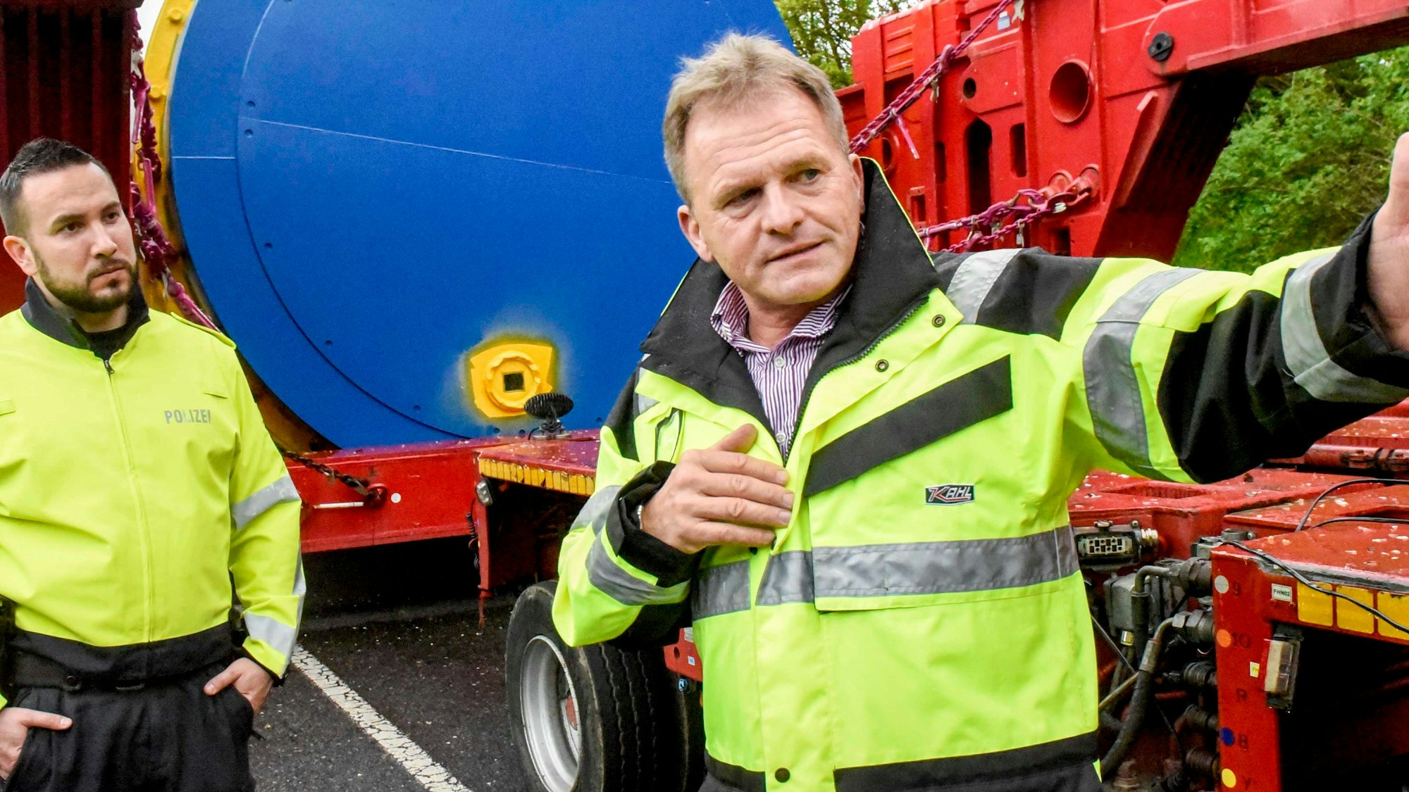 Zwei Männer in gelben Warnjacken stehen vor einem roten Schwertransporter.