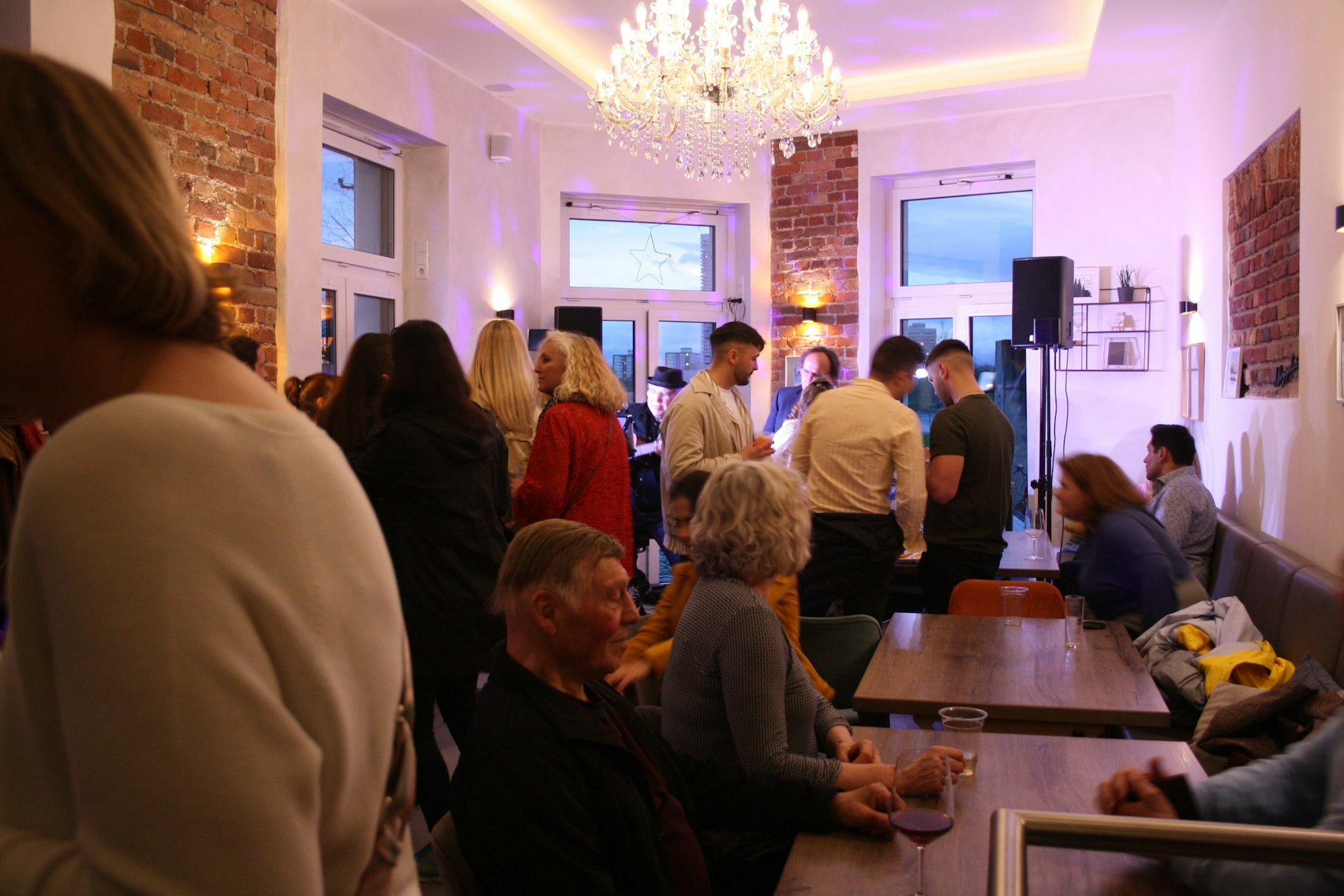 Gäste stehen und sitzen an Tischen in einem hohen Raum, an dessen Decke ein Kristallleuchter hängt.