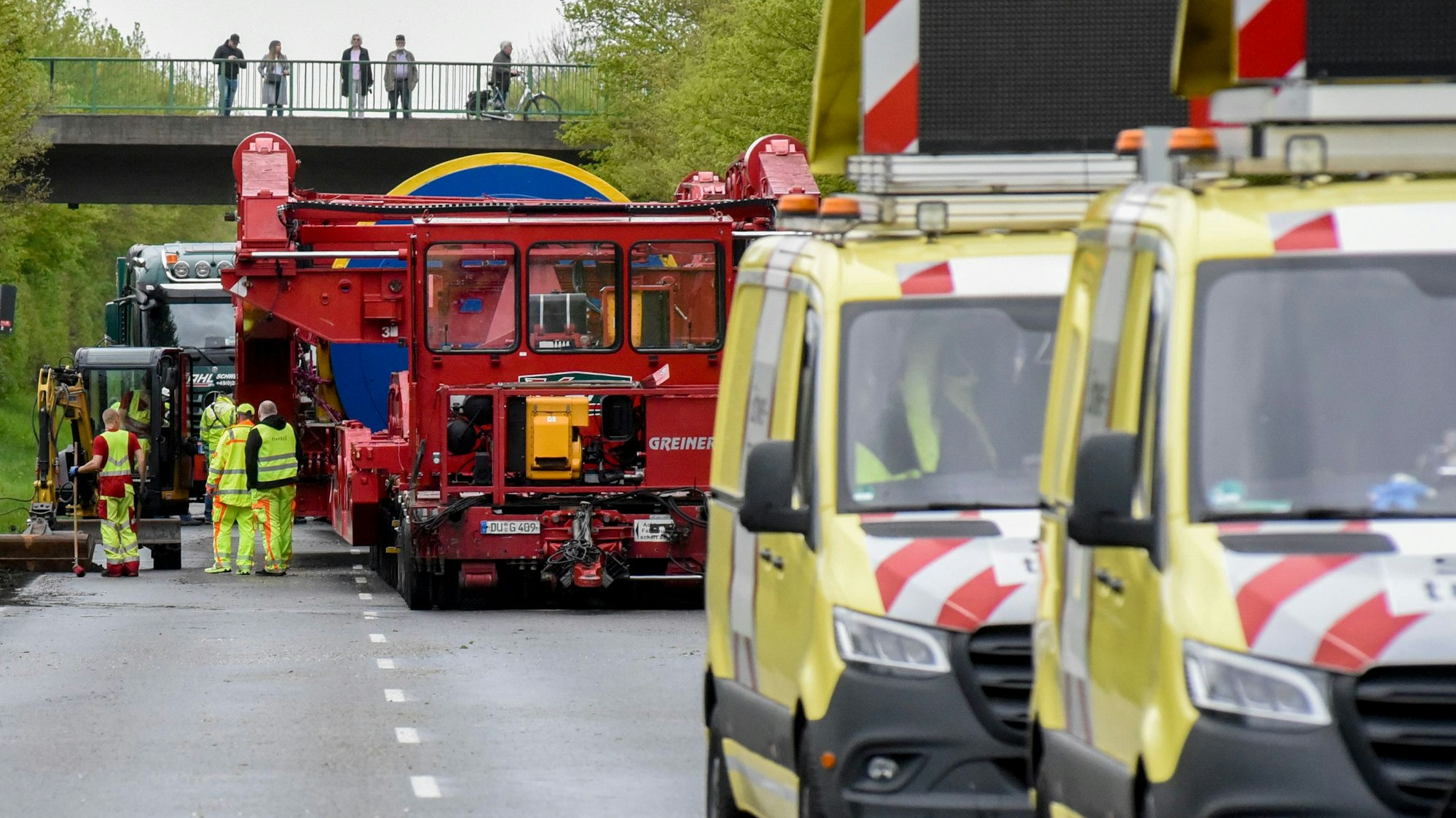 Auf einer Landstraße sind zwei gelbe Fahrzeuge, gefolgt vom roten Schwertransporter, unterwegs. Auf einer Fußgängerbrücke darüber stehen Menschen und sehen zu.