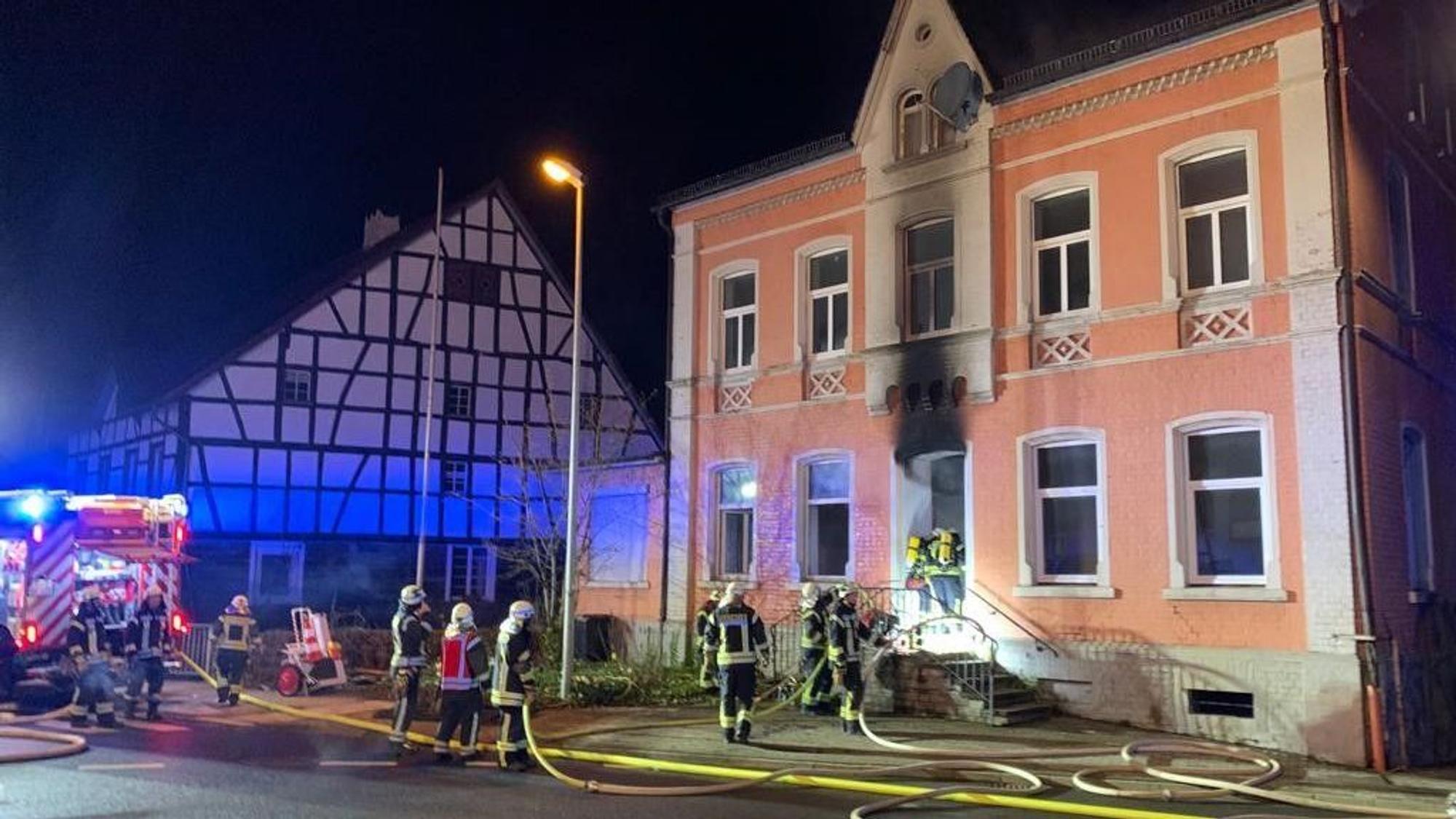 Man sieht die Einsatzkräfte, die in die rosafarbene alte Villa eindringen. Der Ruß des Feuers hat die Fassade über der Eingangstür geschwärzt.Im Kreisverkehr vor dem Gebäude liegen mehrere Schlauchleitungen, die zu einem Feuerwehrfahrzeug führen.