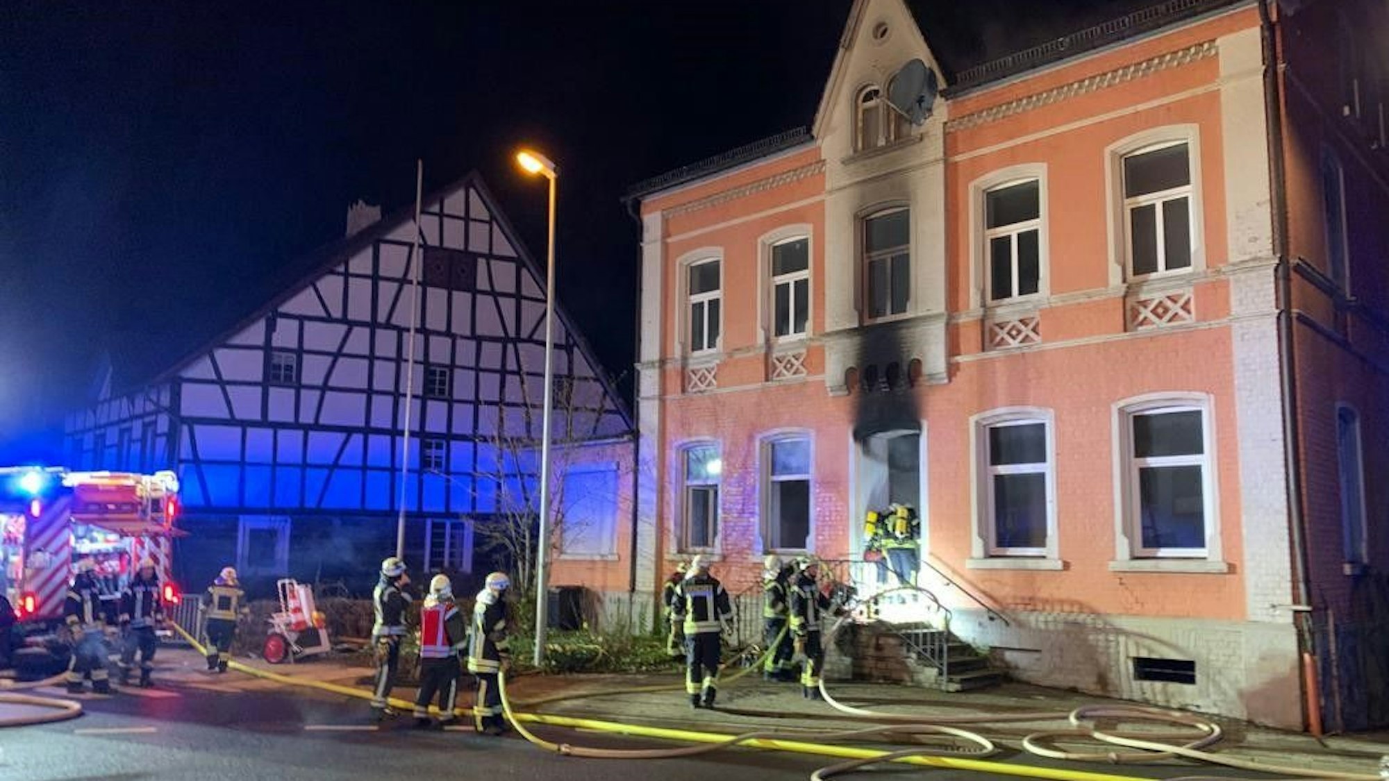 Man sieht die Einsatzkräfte, die in die rosafarbene alte Villa eindringen. Der Ruß des Feuers hat die Fassade über der Eingangstür geschwärzt.Im Kreisverkehr vor dem Gebäude liegen mehrere Schlauchleitungen, die zu einem Feuerwehrfahrzeug führen.