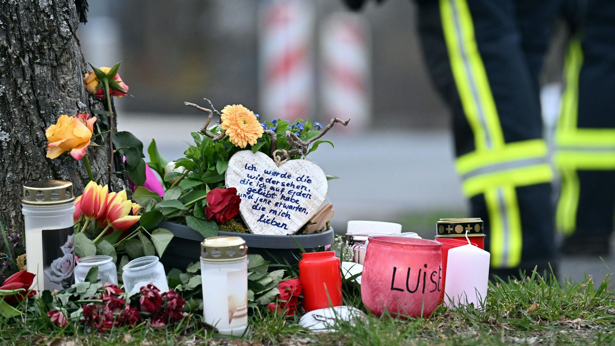 Kerzen und Blumen liegen an einem Baum an der Gesamtschule der getöteten Luise (12) in Freudenberg. Im Hintergrund ist ein Polizist zu sehen.