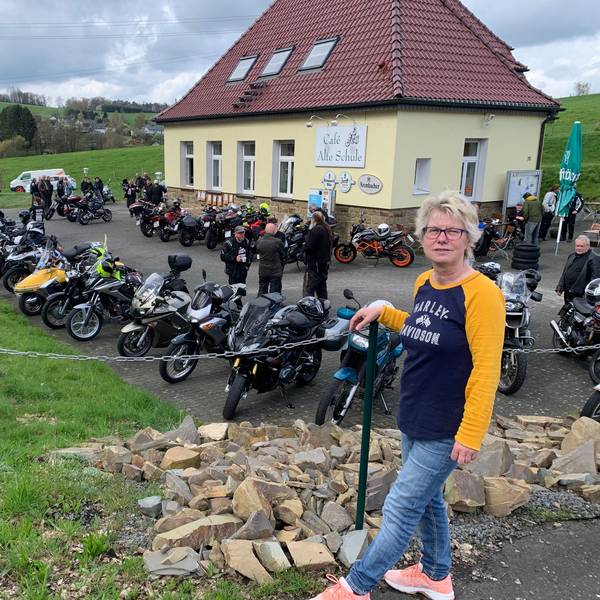 Vor acht Jahren hat Annette Büth die Verantwortung für den Bikertreff übernommen. Eine Frau mit blonden kurzen Haaren steht vor dem Café Alte Schule, auf dem Parkplatz stehen viele Motorräder.