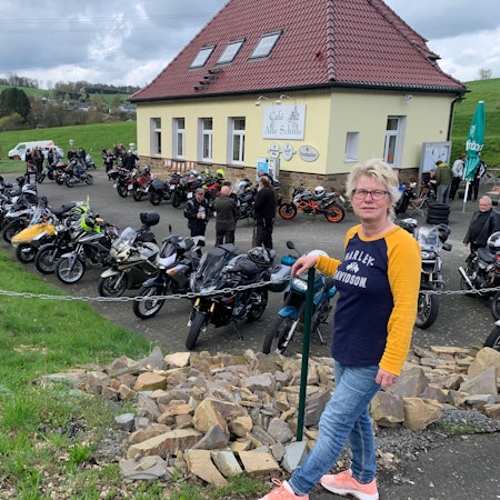 Vor acht Jahren hat Annette Büth die Verantwortung für den Bikertreff übernommen. Eine Frau mit blonden kurzen Haaren steht vor dem Café Alte Schule, auf dem Parkplatz stehen viele Motorräder.