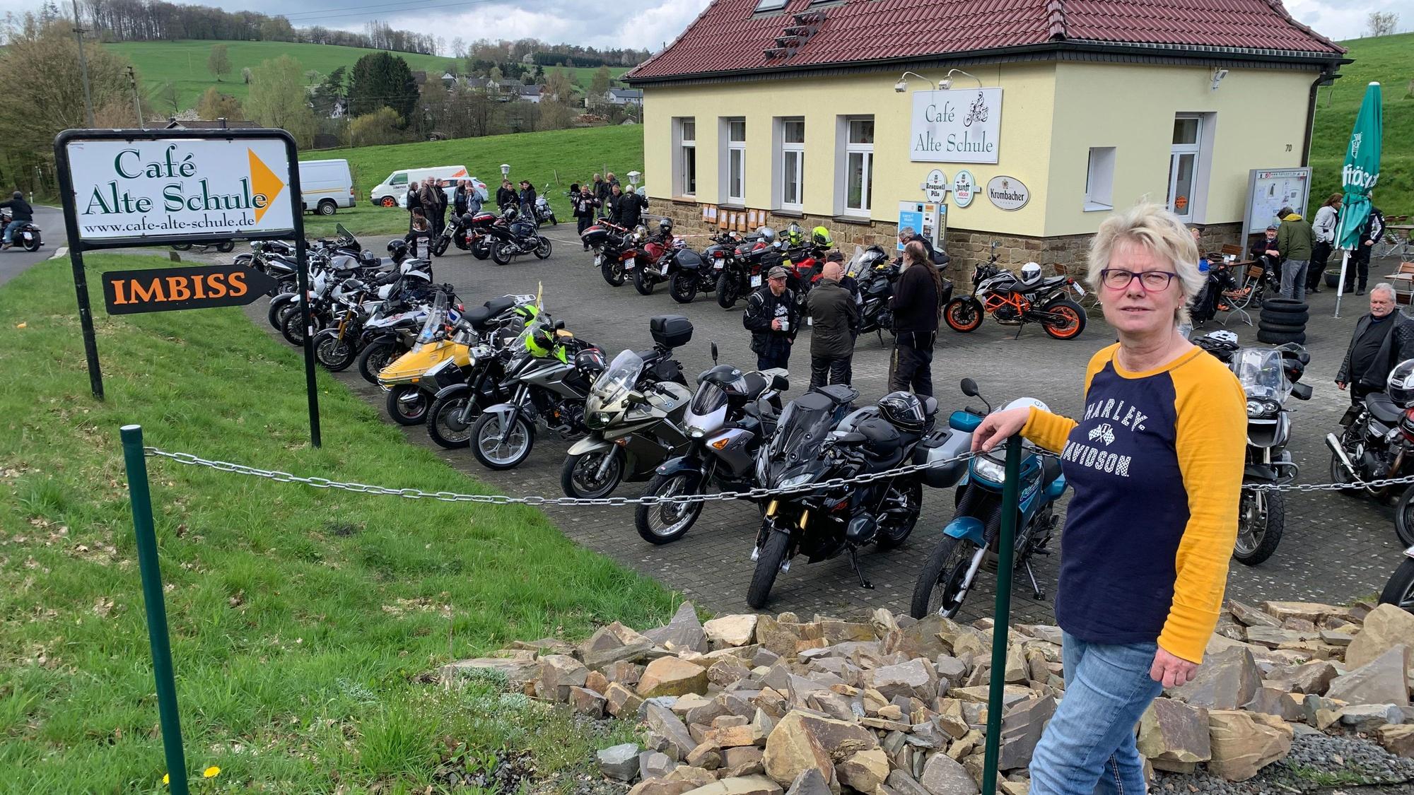 Vor acht Jahren hat Annette Büth die Verantwortung für den Bikertreff übernommen. Eine Frau mit blonden kurzen Haaren steht vor dem Café Alte Schule, auf dem Parkplatz stehen viele Motorräder.