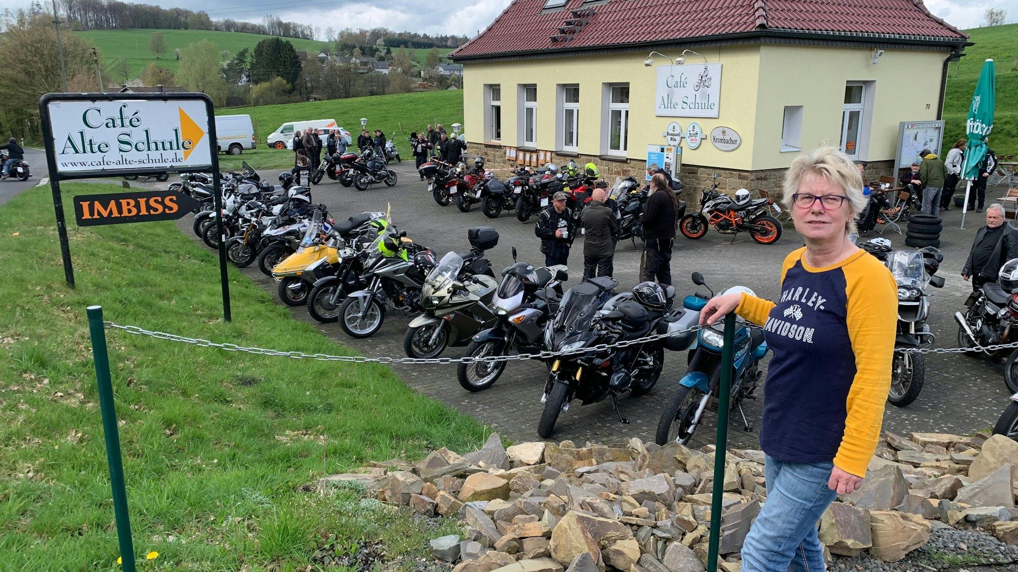 Vor acht Jahren hat Annette Büth die Verantwortung für den Bikertreff übernommen. Eine Frau mit blonden kurzen Haaren steht vor dem Café Alte Schule, auf dem Parkplatz stehen viele Motorräder.