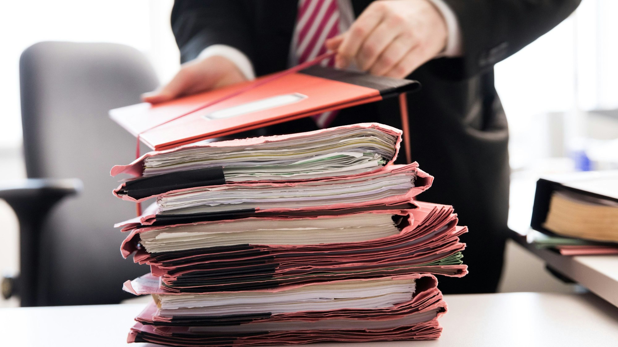 Akten liegen auf einem Schreibtisch. Im Hintergrund steht ein Mann mit Krawatte.