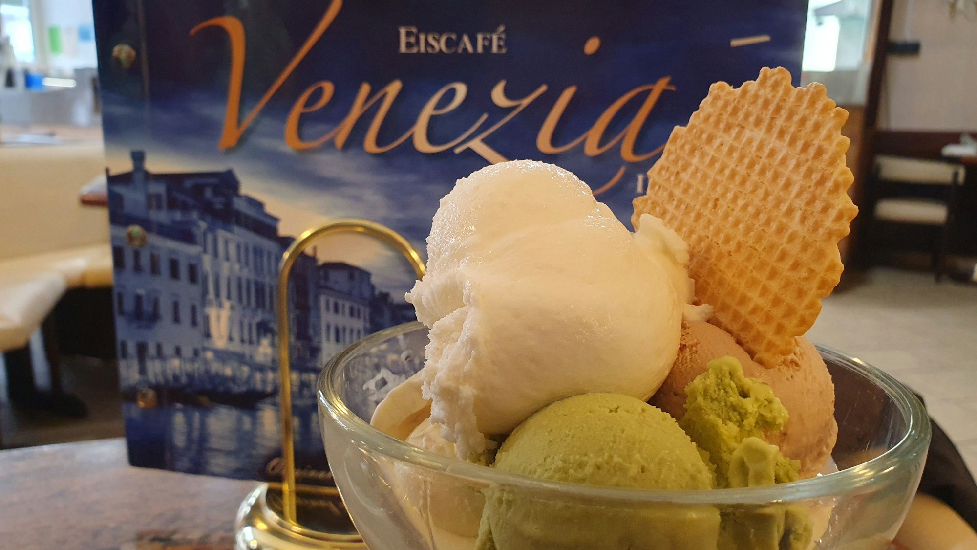 Eisbecher mit vier Kugeln und Waffel vor einer Eiskarte des Eiscafé Venezia.
