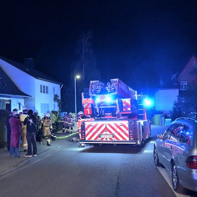 Eine Feuerwehrdrehleiter mit Blaulicht steht in einer Wohnstraße, Feuerwehrleute kommen aus einem Haus. Eine Rettungstrage steht auf dem Bürgersteig.