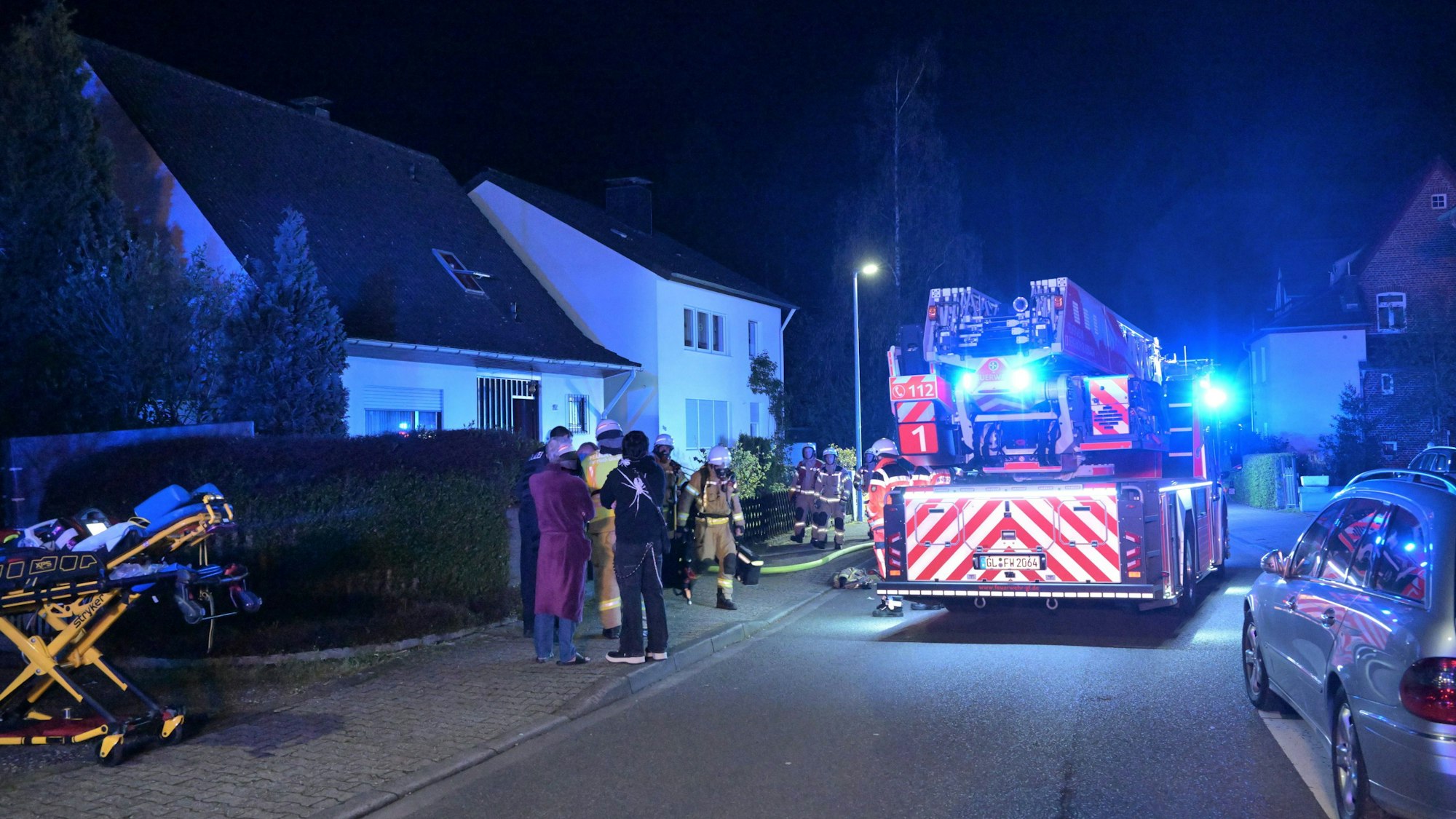 Eine Feuerwehrdrehleiter mit Blaulicht steht in einer Wohnstraße, Feuerwehrleute kommen aus einem Haus. Eine Rettungstrage steht auf dem Bürgersteig.