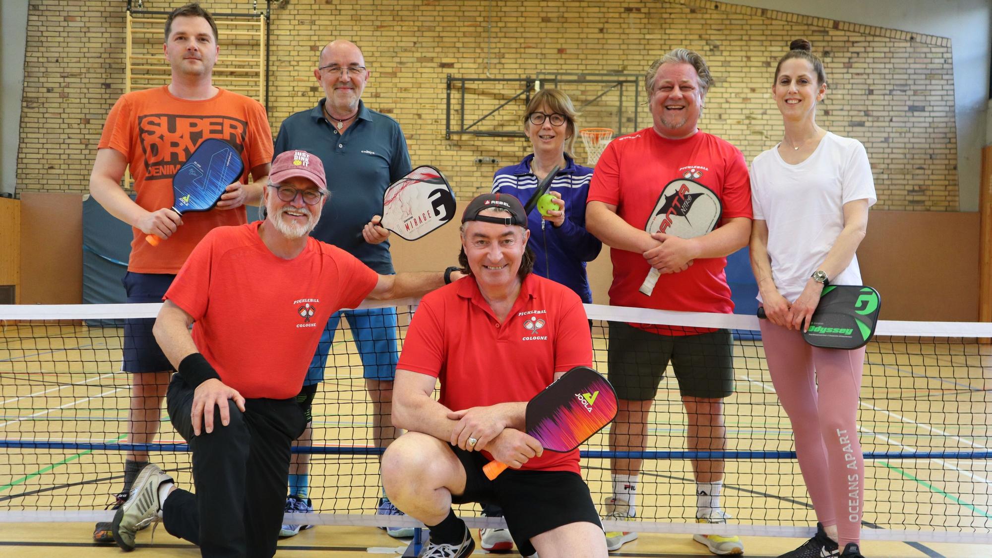 Fünf Männer und zwei Frauen unterschiedlichen Alters in Sportkleidung in einer Turnhalle vor einem Netz mit Schlägern in der Hand