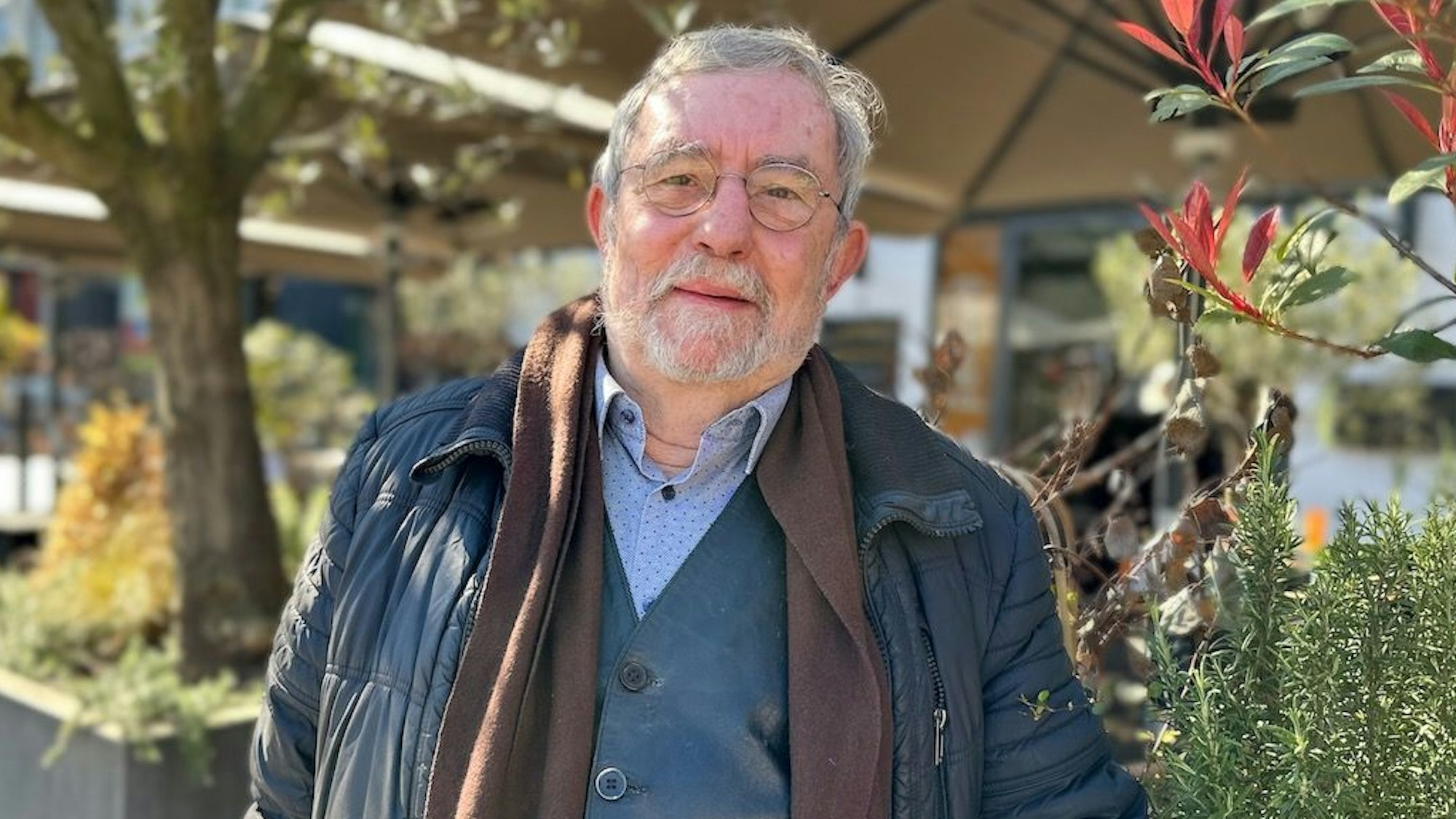 Älterer Mann mit grauen Haaren, Brille, Bart, Schal, Jacket, Hemd und Jacke vor einem Außenbereich mit Pflanzen und Bäumen