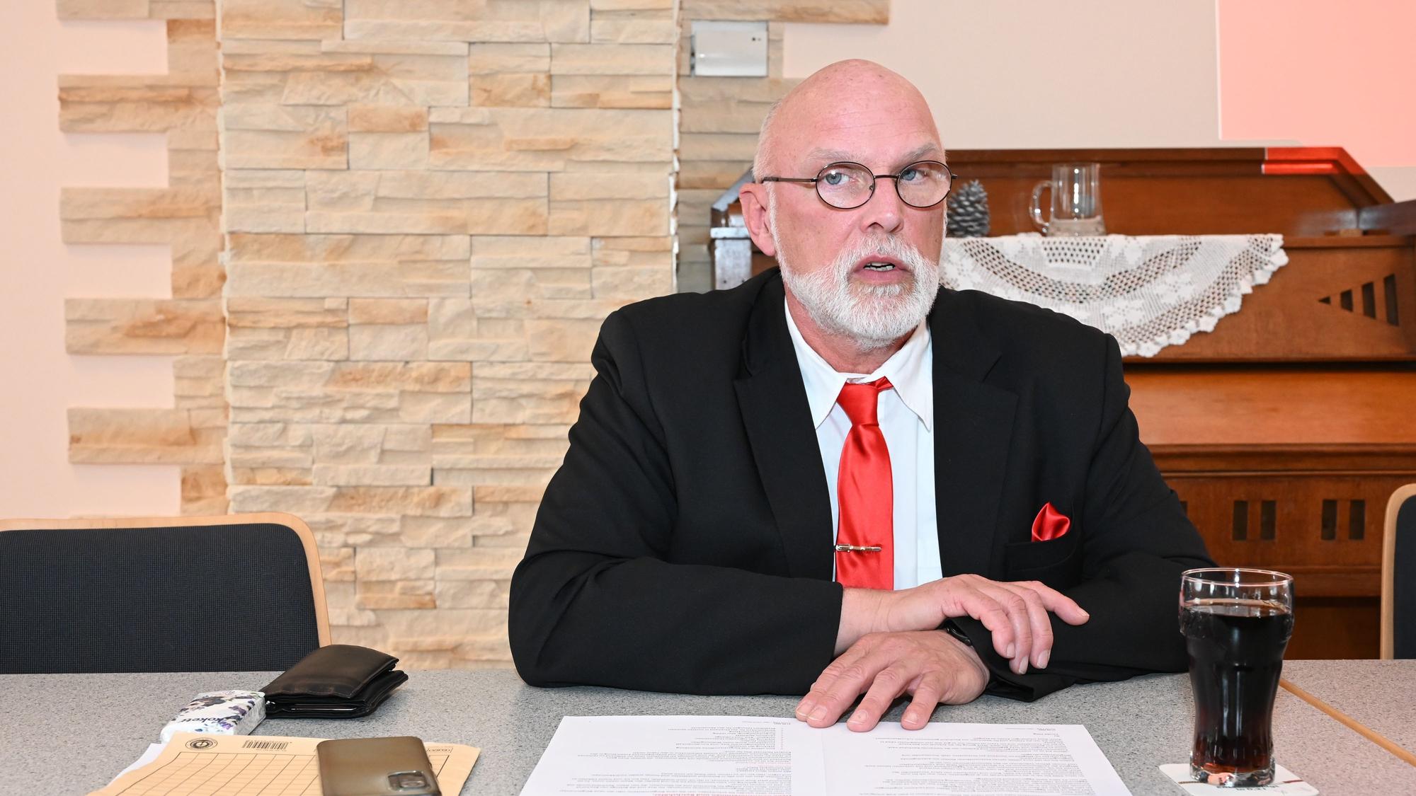 Ein Mann mit Glatze im Anzug und mit roter Krawatte sitzt an einem Schreibtisch.