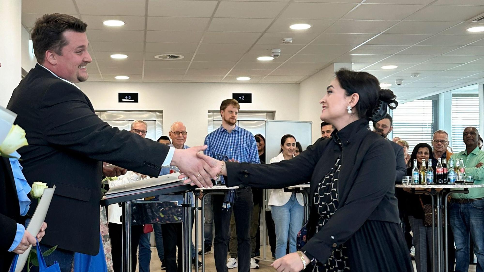 Ein Mann im dunklen Sakko schüttelt einer Frau mit schwarzen Haaren und ebenfalls dunkler Kleidung die Hand. Beide lächeln.