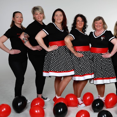 In schwarz-weiß-roten Kleidern posiert das Dreigestirn. Das Gefolge ist komplett in Schwarz angezogen. Auf dem Boden liegen rote und schwarze Luftballons.