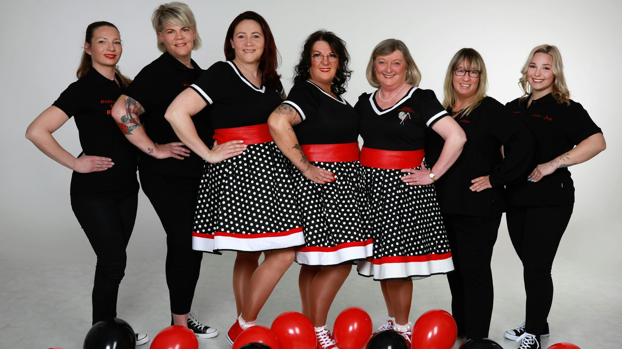 In schwarz-weiß-roten Kleidern posiert das Dreigestirn. Das Gefolge ist komplett in Schwarz angezogen. Auf dem Boden liegen rote und schwarze Luftballons.