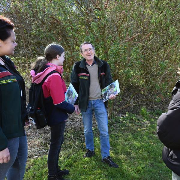 Ein Mann steht mit mehreren Schülerinnen im Wald. Er hat eine Informationsmappe in der Hand und spricht zu den jungen Frauen.