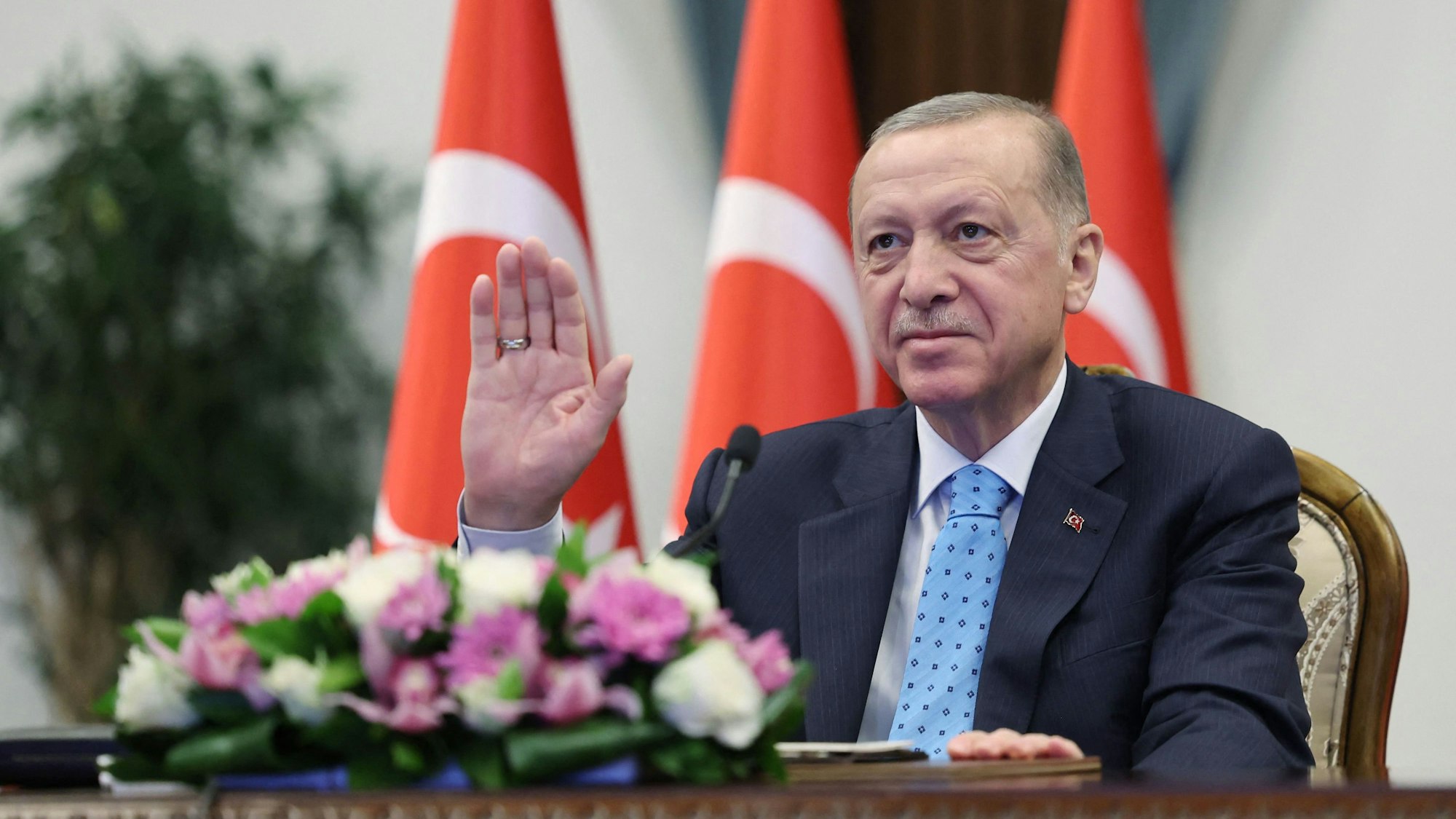 Recep Tayyip Erdogan sitzt an einem Tisch und hebt die rechte Hand.