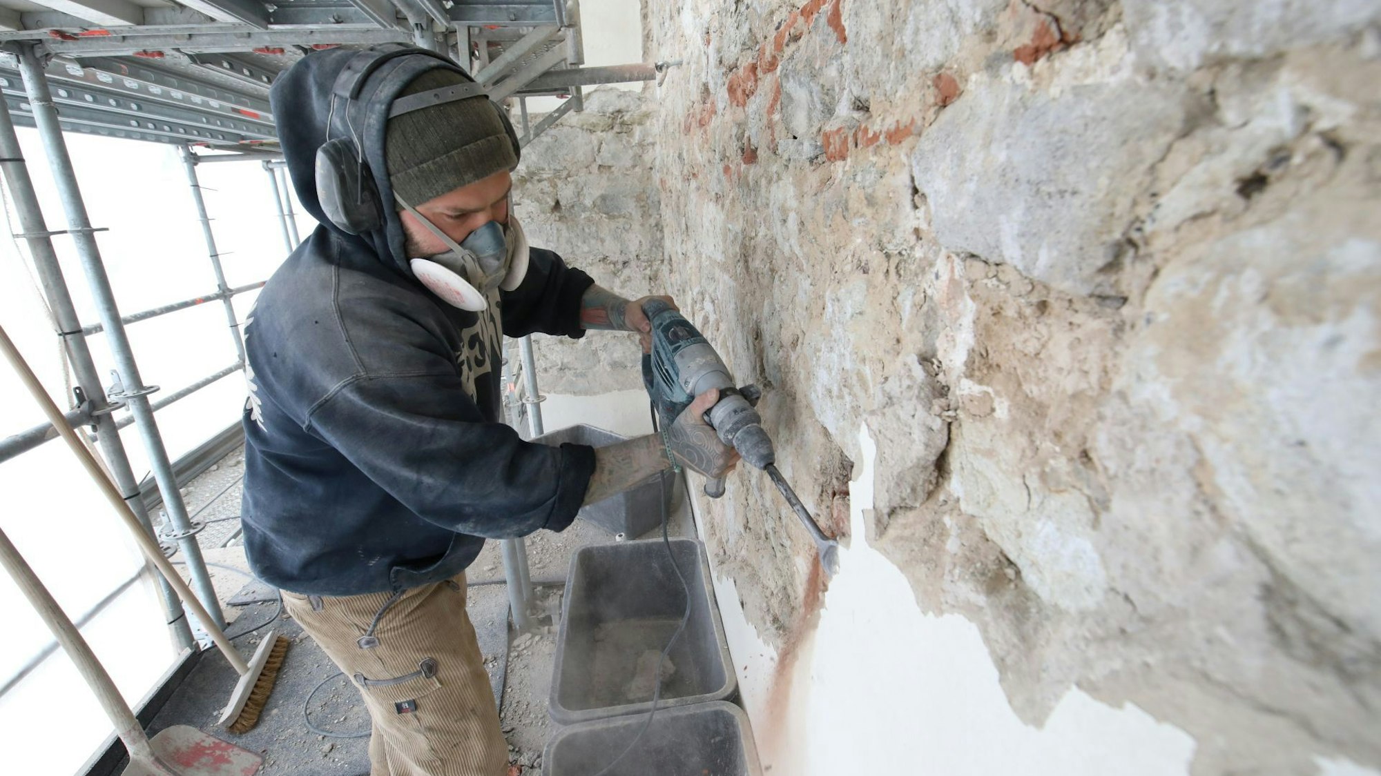Ein Arbeiter steht auf dem Gerüst und trägt den Putz mit einem Bohrer ab.