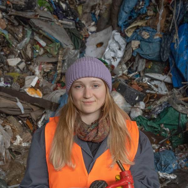 Eine Studentin der TH Köln steht vor einem Berg aus Müll. Sie trägt eine lila Mütze und eine orange Warnweste.