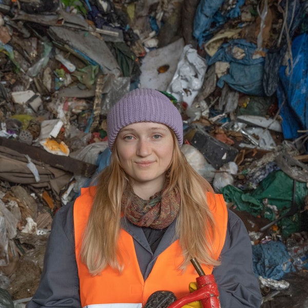 Eine Studentin der TH Köln steht vor einem Berg aus Müll. Sie trägt eine lila Mütze und eine orange Warnweste.