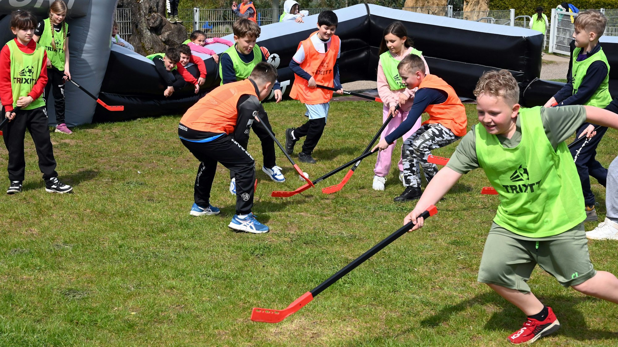 Kinder in orangefarbenen und gelben Leibchen spielen auf einem Feld auf einer Wiese Hockey.