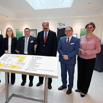 Mehrere Männer und Frauen stehen in einem Gerichtsgebäude vor einer großen Tafel für Sehbehinderte.