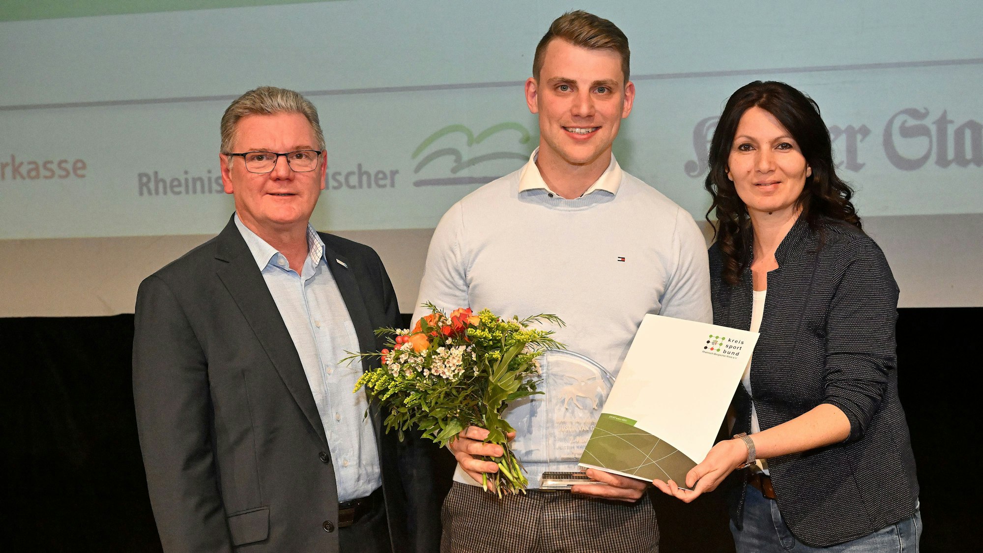 Drei Menschen stehen mit Blumen und Sportlerehrungsurkunge auf einer Bühne.